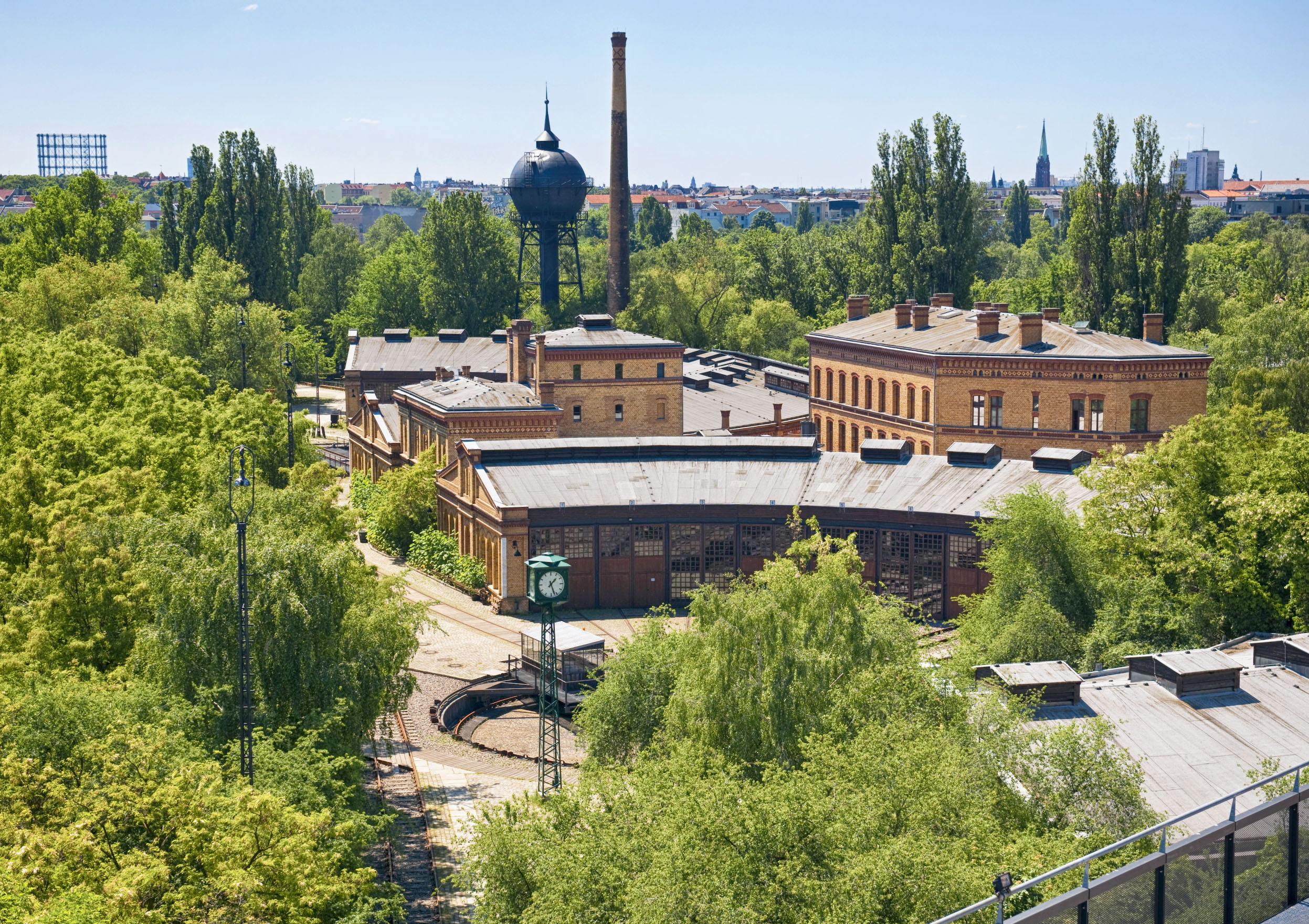 A bird's-eye view of a group of brick buildings surrounded by green trees. Behind the buildings are a water tower and a chimney.