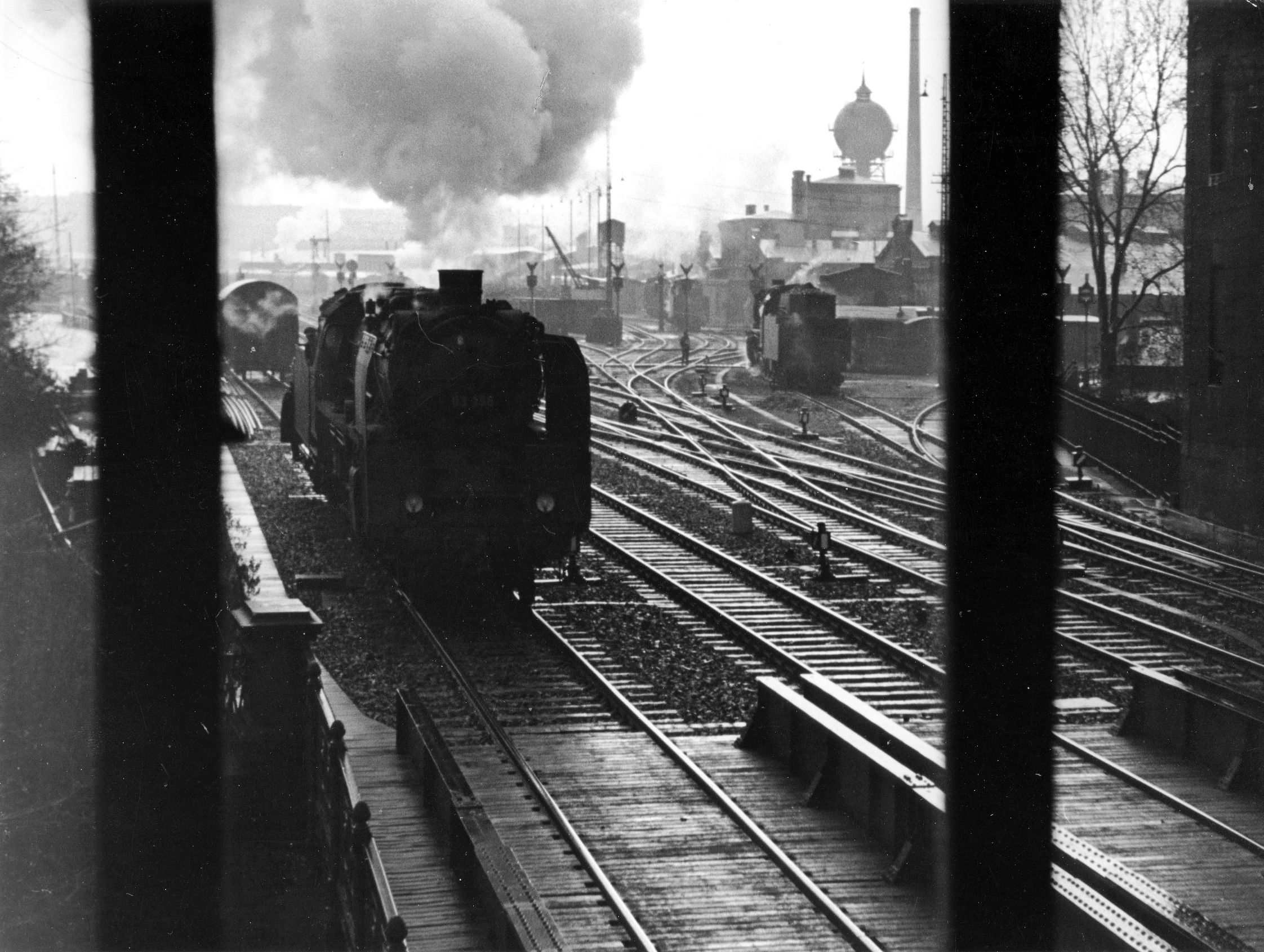A black-and-white photo of a steam locomotive entering the frame on a track. A water tower and a locomotive shed can be seen in the background.