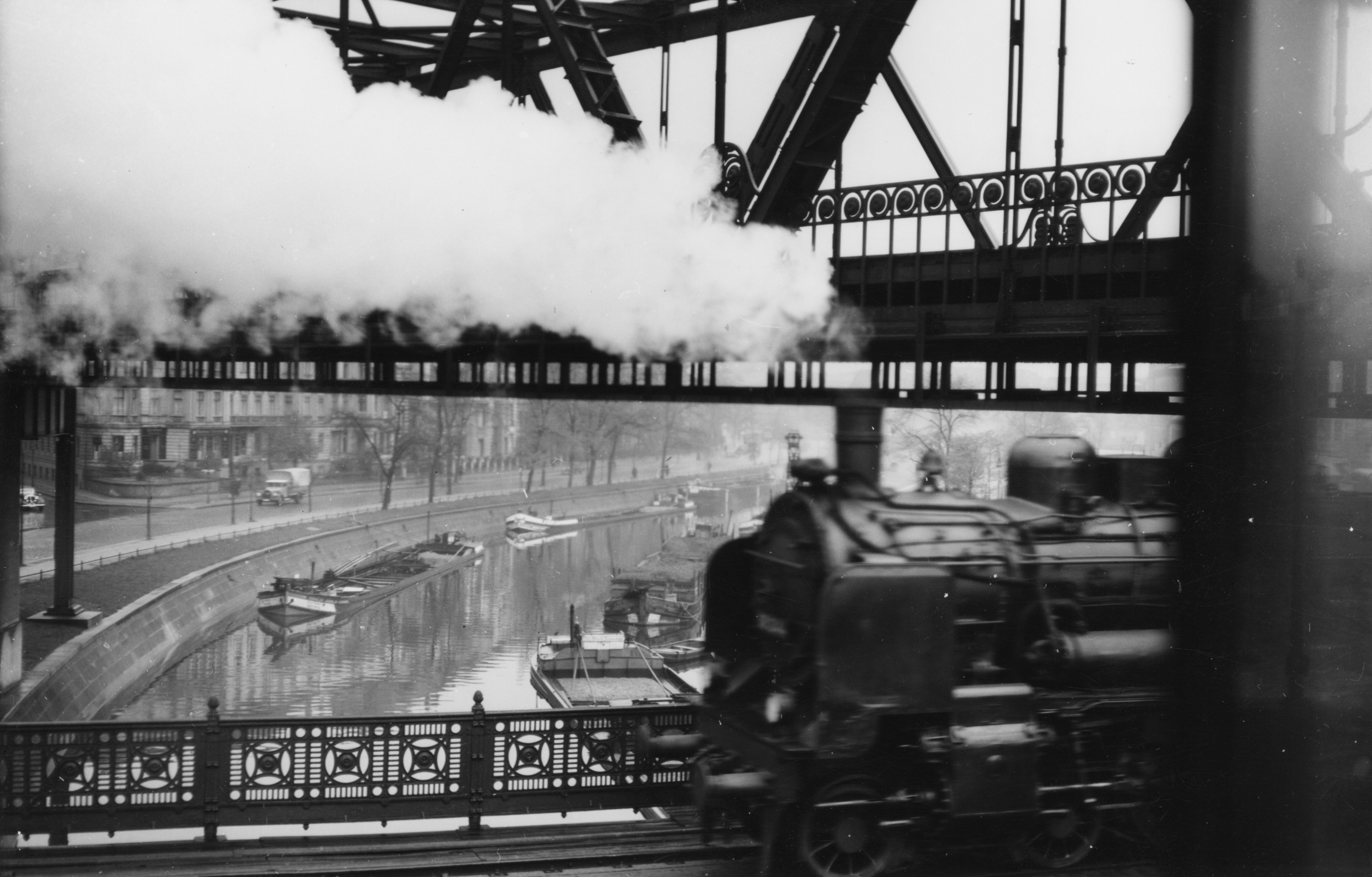 A black-and-white photo of a locomotive shrouded in clouds of steam as it crosses a canal on a bridge.