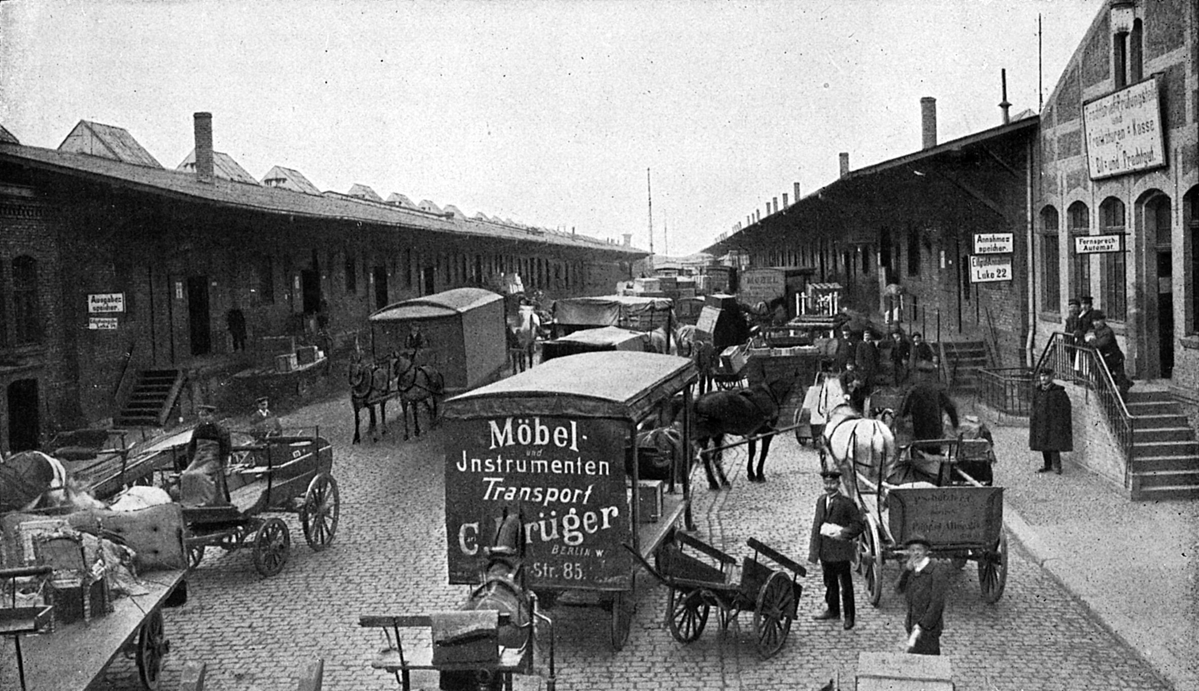 A black-and-white photograph of a cobblestone street lined with warehouses on both sides. Horse-drawn carts, wagons, and people are moving along the street.