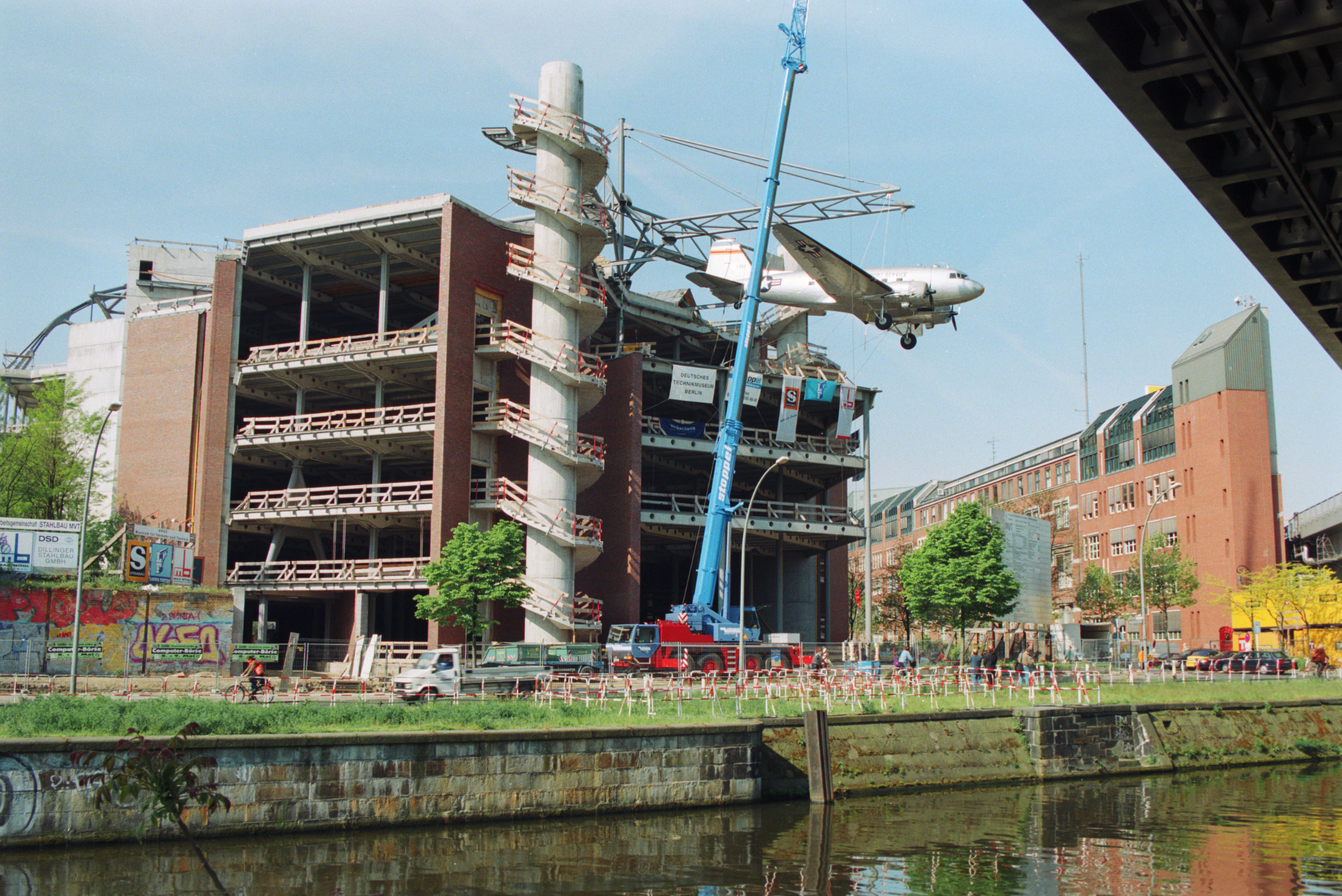 The shell of a four-story building. An airplane is suspended from a steel structure on the roof. A blue crane stands in front of the building.