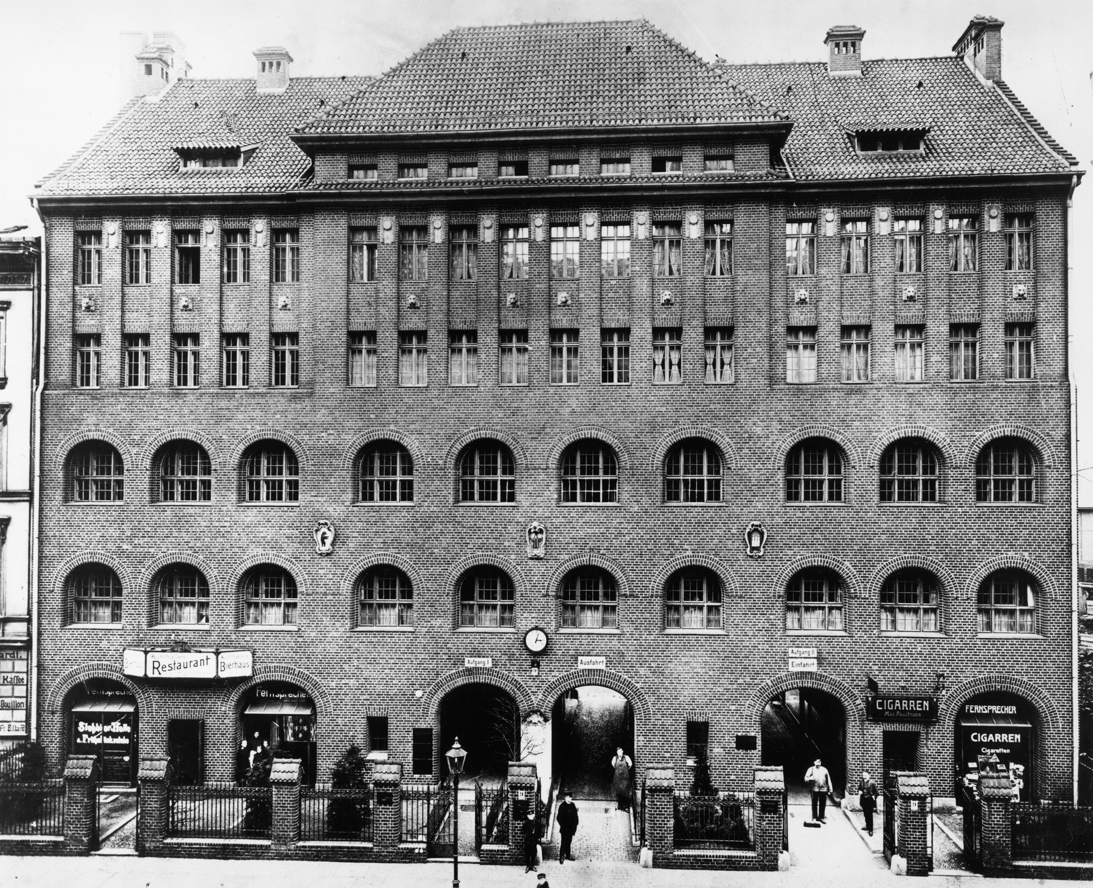 Black-and-white photo: A front view of a large, four-story brick building. On the ground floor, large arched doorways lead into the building.