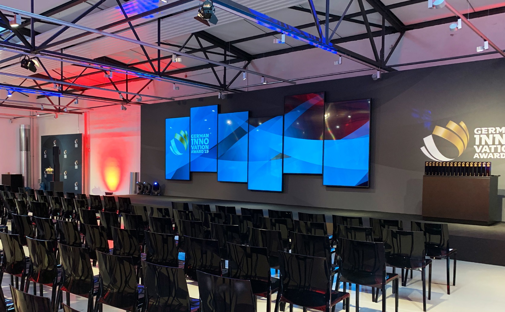 In einer Halle sind Stuhlreihen und ein Podium mit Monitoren an der Rückwand zu sehen.
